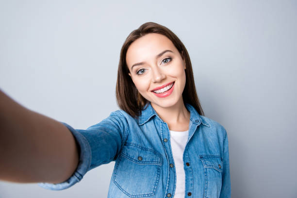 selfie manie. nahaufnahme von zuversichtlich brünette mädchen mit strahlenden lächeln in denim oufit grauen hintergrund ein selbstporträt auf ihr handy und ständigen übernahme - selfie fotos stock-fotos und bilder