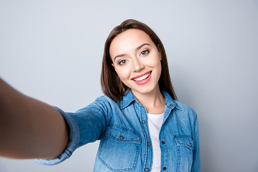 Selfie mania. Close up of confident brunette girl  with beaming smile in denim oufit taking a self-portrait  on her mobilephone and  standing over grey background