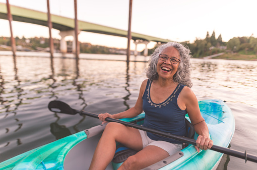 Ethnic mature female kayaks in river on a summer evening. She has a huge smile and is looking at the camera. There is a bridge behind her.