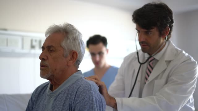 Concerned doctor checking the lungs of his patient with a stethoscope while dictating something to the nurse