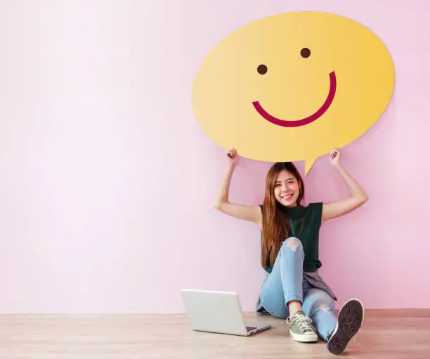 Photo of Happy Customer Concept. Review and Feedback her Experience for Satisfaction Survey Online. Young Female in Cheerful Posture, Raise up Speech Bubble with Smiley Face. Sit on the Floor with Laptop