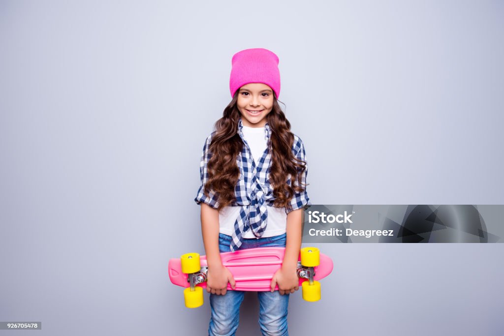 Portrait of little cute smiling girl with pink skate-board in pink hat who is ready to ride on the street standing over grey background Child Stock Photo