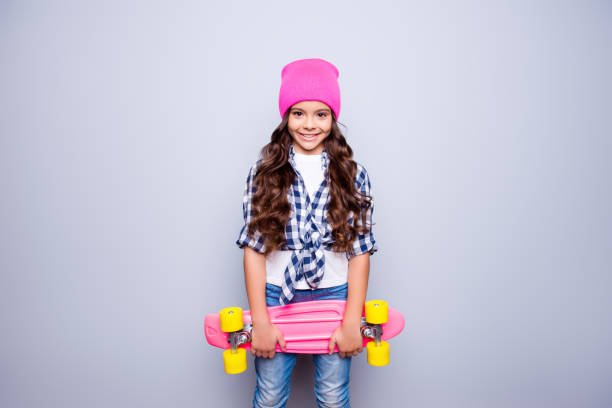 portret małej uroczej uśmiechniętej dziewczyny z różową deskorolką w różowym kapeluszu, która jest gotowa do jazdy na ulicy stojącej nad szarym tłem - fashion male beauty confidence elegance zdjęcia i obrazy z banku zdjęć