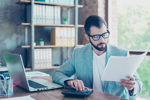 Portrait of young, serious accountant holding papers in hand and sum up accounts of the company for last month while sitting at his desktop in the workstation