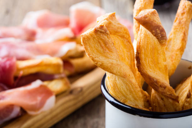 Bread sticks with cheese puff pastry in rustic white mug Bread sticks with cheese puff pastry in rustic white mug on cured ham and glass of red wine background on wooden table, delicious snack twisted bacon stock pictures, royalty-free photos & images