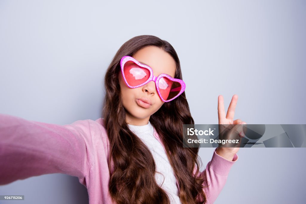 Autorretrato de muy niña haciendo selfie gesticular símbolo de la paz en ropa casual y gafas en forma de corazón mientras que se coloca sobre el fondo gris de cerca - Foto de stock de Autofoto libre de derechos