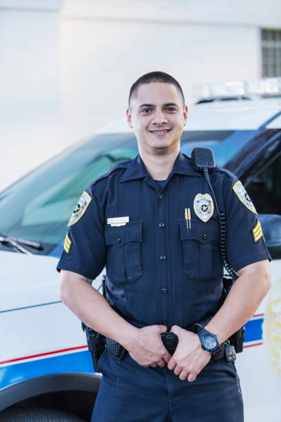 Hispanic police officer standing by squad car An Hispanic policeman standing in front of his police car. The officer in uniform is a mid adult man in his 30s, smiling confidently at the camera. police force smiling officer stock pictures, royalty-free photos & images