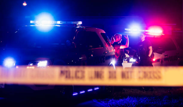 Police officers in bulletproof vests behind cordon tape Two multi-ethnic police officers at night wearing bulletproof vests, standing beside police cars with emergency lights flashing, behind cordon tape. The policewoman is in her 40s and her Hispanic, male partner is in his 30s. police vehicle lighting stock pictures, royalty-free photos & images