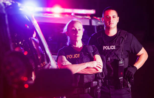 Police officers in bulletproof vests at night Two multi-ethnic police officers at night wearing bulletproof vests, standing beside police cars with emergency lights flashing. The policewoman is in her 40s and her Hispanic, male partner is in his 30s. police car photos stock pictures, royalty-free photos & images