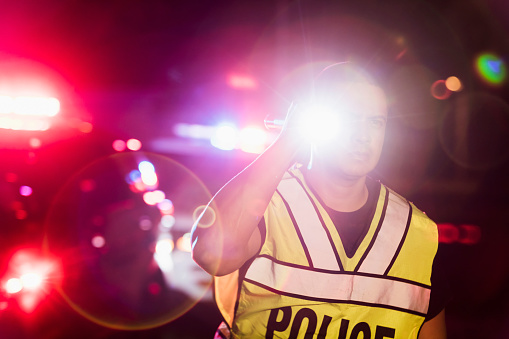 An Hispanic police officer wearing a reflective vest, standing in front of police cars with emergency lights flashing, looking away with a serious expression, holding a flashlight. He is a mid adult man in his 30s.