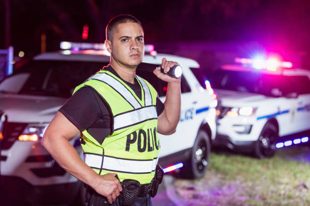 An Hispanic police officer wearing a reflective vest, standing in front of police cars with emergency lights flashing, looking away with a serious expression, holding a flashlight and resting a hand on his holster. He is a mid adult man in his 30s.