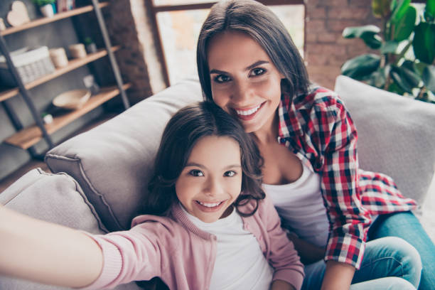 schön mit toothy lächeln mutter und ihr niedliche tochter sind mit einer video-unterhaltung mit dem vater, der jetzt im ausland ist, sitzen sie auf einem sofa zu hause - offspring child toothy smile beautiful stock-fotos und bilder