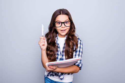 Portrait of little, brunette, cute, smiling girl holding notebook raising her pencil up, finding a solution how to do exercise, standing over grey background