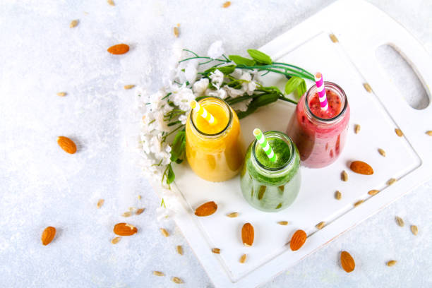 Green, yellow, purple smoothies in bottles of berries, greens, oranges on a white board on a gray table.