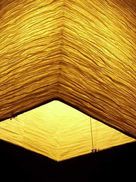 Rectangular Asian hanging lamp seen from below, made of crinkled rice or mulberry paper.  Shallow DOF with focus on opposite side and sides. Black copy space is left at the bottom.  DSLR Photograph is in vertical format and used a low ISO to avoid noise in the shadows. Lamp is glowing in the blackness.