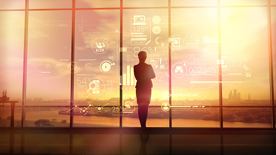 Silhouette of a woman in a huge office in front of large windows viewing corporate infographics.