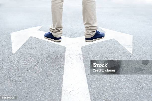 Young Adult Standing On Arrows To Make Decision Stock Photo - Download Image Now - Crossroad, Forked Road, Arrow Symbol