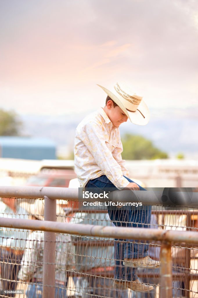 Little Buckaroo Cowboy Sitting On A Tall Fence Smiling To Himself As The Adults All Gather and Talk Down Below 6-7 Years Stock Photo