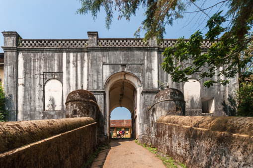 Madikeri, India - October 31, 2013: Gray-white stone entrance to royal fort. Walkway over moat bridge. Green vegetation. Blue sky. Some people seen through gate.