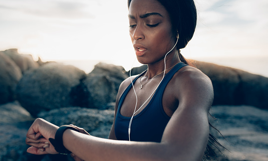 Close up of young fitness woman looking at her smart watch while taking a break from outdoor workout. Sportswoman checking pulse on fitness smart watch device.