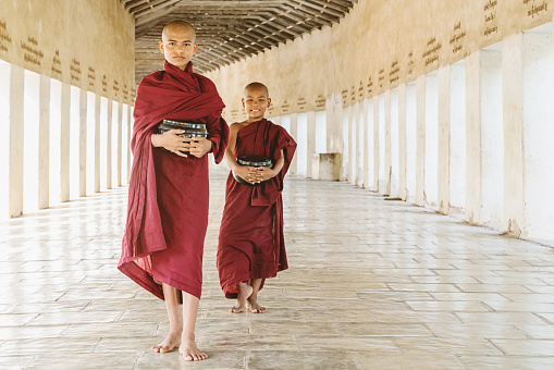 Burmese buddhist novice monks in their typical religious veils walking together along monastery archway with their alms bowls in their hands. Bagan, Mandalay Region, Myanmar.