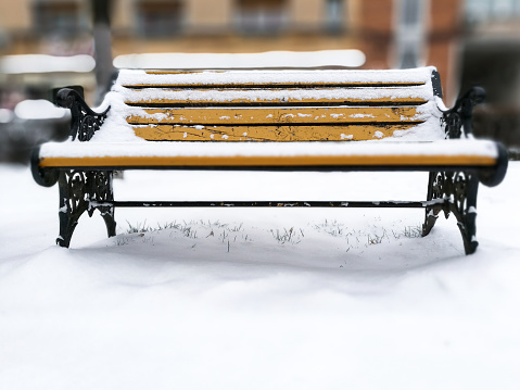 Close up image depicting a bright yellow park bench covered in snow. The ground around the bench is similarly covered in snow. The background is blurred out of focus leaving lots of room for copy space. Images taken on mobile device.