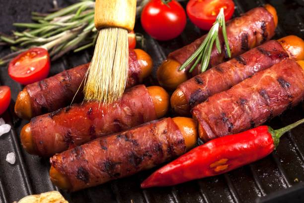 праздничные коктейльные сосиски, завернутые в хрустящий копченый бекон, широко известный как «свиньи в бланкетах» в черной кастрюле - blanket pig hot dog sausage стоковые фото и изображения