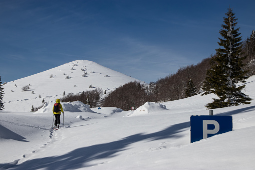 Woman on skis walking past parking road to snow covered mountain peak