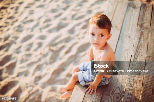 The Theme Is A Child And Summer Beach Vacation A Small Caucasian Boy Sits Sideways On A Wooden Pier And Looks At The Camera On A Sandy Beach And A Pond A River With Bare Legs In Blue Denim Shorts Stock Photo - Download Image Now