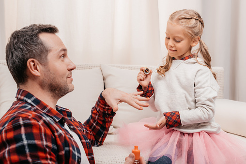cute little girl polishing nails of smiling father at home