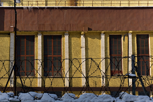 sunlit wall of an industrial building behind a snow-covered fence with barbed wire