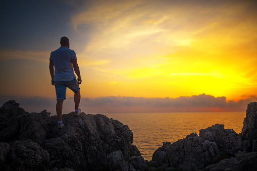 Man stands on the rocks at the sea and enjoys a sunset