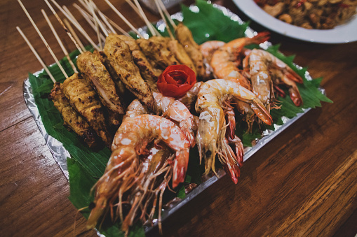 Asian Thai snacks are located on a plate standing on a wooden table. There are shashliks on skewers, fried shrimps and vegetables. The plate is lined with green leaves. Close up.