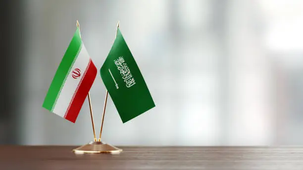 Iranian and Saudi Arabian flag pair on desk over defocused background. Horizontal composition with copy space and selective focus.