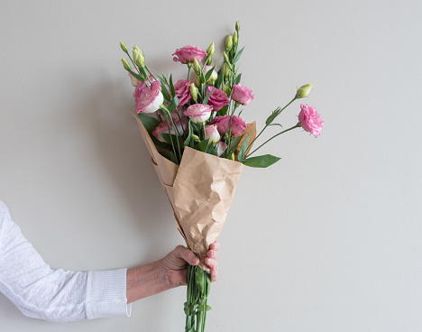 Woman's hand holding bouquet of pink lisianthus flowers wrapped in in brown paper against neutral wall background