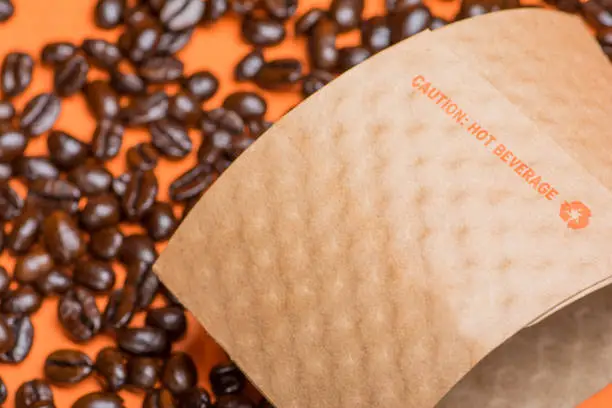 Brown paper cup holder used on coffee cups to protect the hands from heat. Whole coffee beans on orange background.