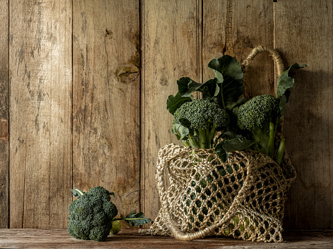 Fresh market broccoli in a pretty hemp string reusable shopping bag against an old wood paneled wall with other broccoli sprouts on a table beside the bag.