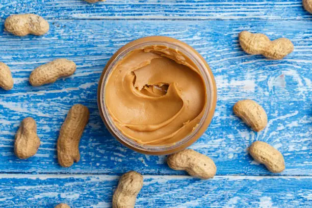 peanut butter in an open jar on a blue wooden background, next to peanuts scattered