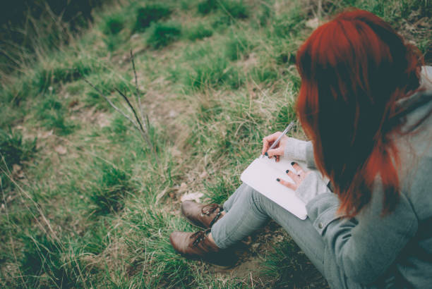 рыжая девушка сидит на зеленой весенней траве, пишущей в блокноте. - writing diary nature ideas стоковые фото и изображения