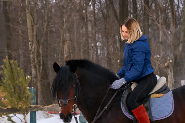 A young girl with white hair learns to ride a horse. The girl recently started to practice equestrianism. The girl is afraid of riding a horse quickly. A cloudy winter day."n