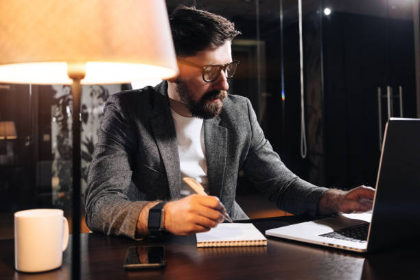 istock Bearded project manager hold pen in hand and using contemporary notebook. Young businessman working in loft office at night. Man making new startup 927502542