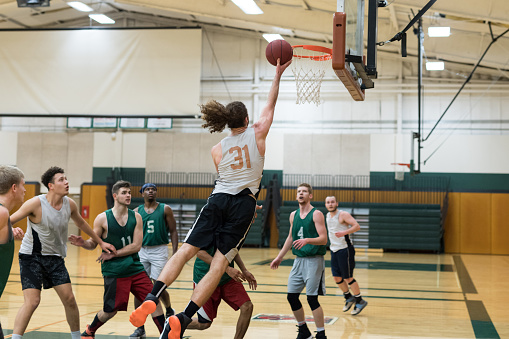 A men's college basketball team practices in the gym. A player is leaping into the air for a layup and about to score as others watch and box each other out.