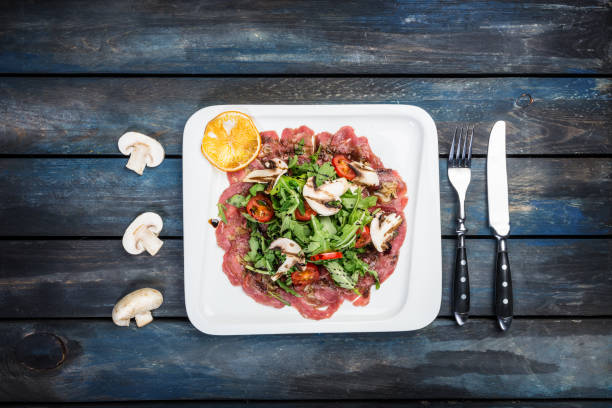 Beef carpaccio with mushrooms on wooden background served with cutlery Marinated beef stuffed porcini mushrooms with a salad of arugula with balsamic dressing, top view carpaccio parmesan cheese beef raw stock pictures, royalty-free photos & images