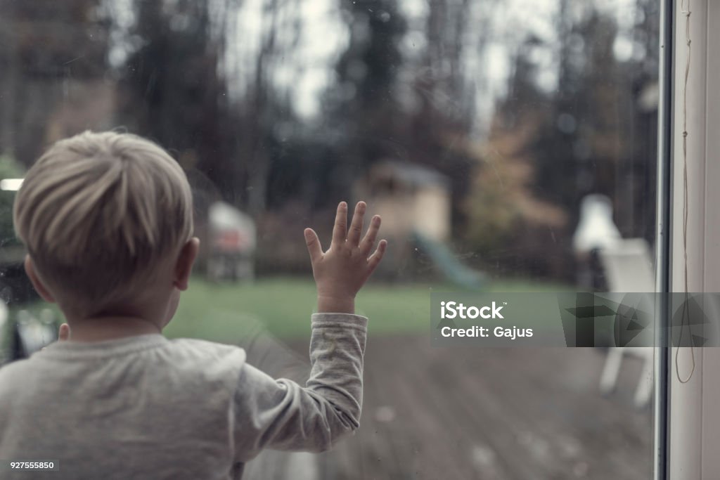Toddler boy staring out of a window in evening light into the garden Toddler boy staring out of a window in evening light into the garden with his hand spread out on the glass, toned retro effect. Child Stock Photo