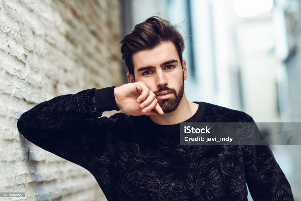 Young bearded man, model of fashion, in urban background wearing casual clothes. Young bearded man, model of fashion, looking at camera in urban background wearing casual clothes. Guy with beard and modern hairstyle in the street. Adult Stock Photo