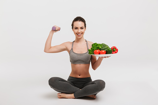 happy young woman sitting on floor with tray of various healthy vegetables and showing her muscles isolated on white