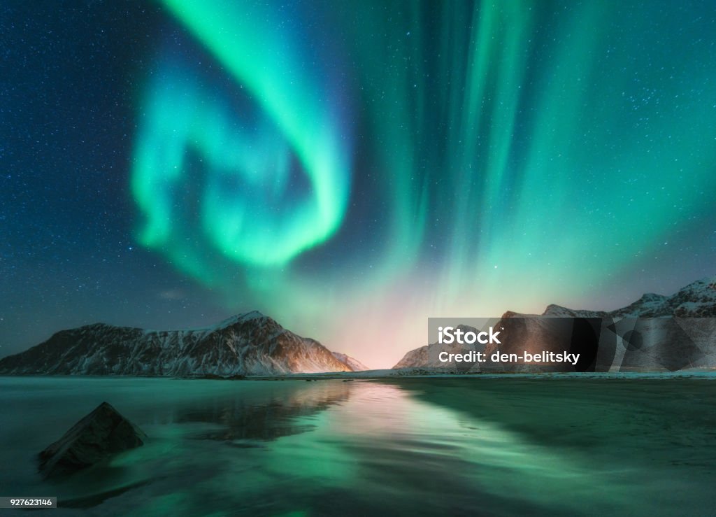 Aurora borealis in Lofoten islands, Norway. Aurora. Green northern lights. Starry sky with polar lights. Night winter landscape with aurora, sea with sky reflection, stones, beach and snowy mountains Aurora Borealis Stock Photo