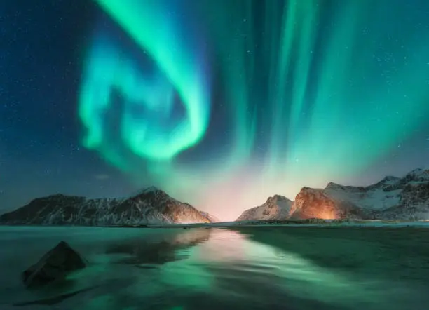 Aurora borealis in Lofoten islands, Norway. Aurora. Green northern lights. Starry sky with polar lights. Night winter landscape with aurora, sea with sky reflection, stones, beach and snowy mountains