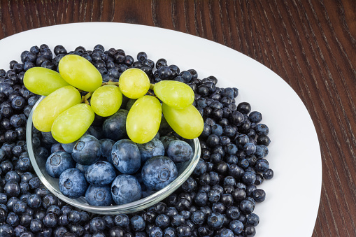Close-up of white plate with luscious fresh berry fruits and glass bowl on wooden background.