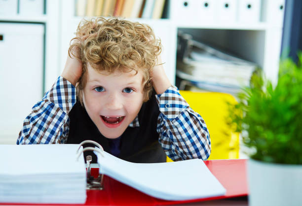 istock Small Caucasian curly boy grabbed his head while sitting over documents in the office. The boy plays in an office worker. 927641452